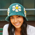 Woman wearing a teal crochet hat with floral decorations, sitting outdoors.
