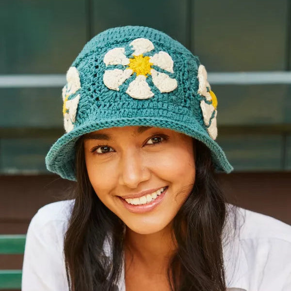 Woman wearing a teal crochet hat with floral decorations, sitting outdoors.