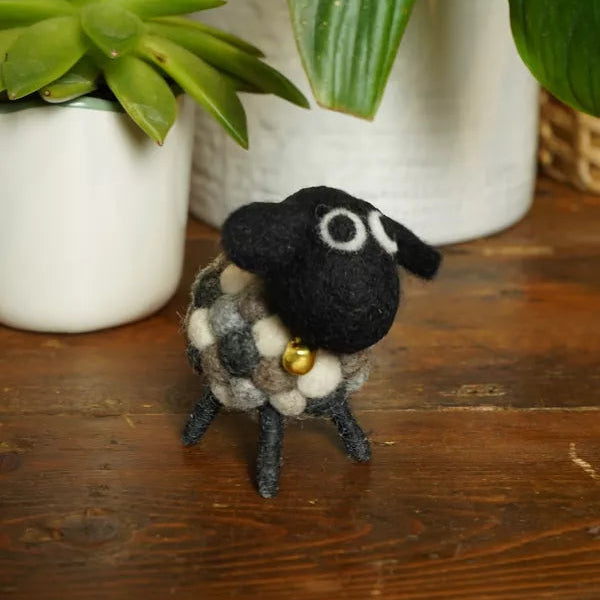 Small black and white sheep figurine with a gold bell on a wooden surface with plants in the background.