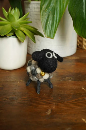 Small black and white sheep figurine with a gold bell on a wooden surface with plants in the background.