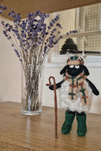 Toy sheep figure with a scarf and walking stick next to a vase of lavender on a wooden surface.