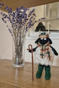 Toy sheep figure with a scarf and walking stick next to a vase of lavender on a wooden surface.