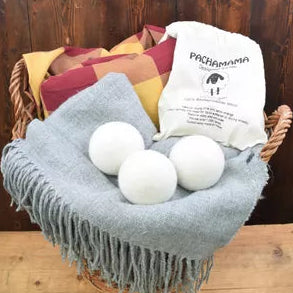 A gray fringed throw blanket draped over a wicker basket, with three white spherical bath balls resting on top and a white fabric pouch labeled 'PACHAMAMA' nearby. The basket is set against a dark wood plank background.