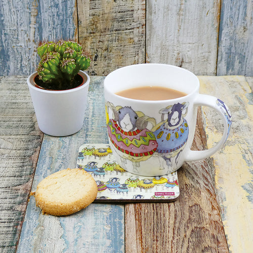 A white ceramic mug with a pattern of colorful sheep wearing sweaters, placed on a wooden surface next to a small potted plant and a cookie.